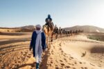 Travellers walking with a camel caravan through the Sahara dunes